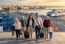 Photographie aérienne d'une famille multigénérationnelle de la diaspora (6 personnes) débarquant joyeusement d'un ferry dans le port de Tunis (La Goulette). Au premier plan, un couple qualifié tire des valises, un grand-parent est ému, et un enfant tient un drapeau tunisien. En arrière-plan, le grand ferry 'TUNISIA LINE' et les infrastructures portuaires sont éclairés par la lumière dorée du lever du soleil.