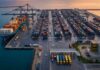 Photographie éditoriale aérienne du port de Radès en Tunisie au lever du soleil. Des portiques de déchargement modernes, des piles de conteneurs et un navire cargo sont visibles.